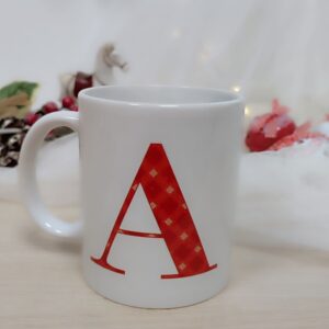 Taza de cerámica blanca decorada con inicial en una cara y bouquet navideño en la otra.